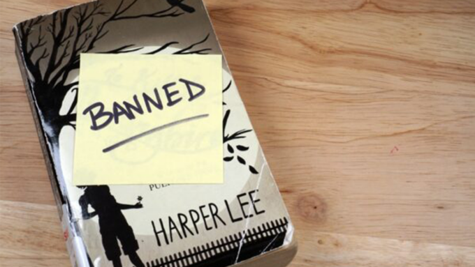 Harper Lee's To Kill a Mockingbird on a wooden table with a yellow sticky note reading BANNED on the cover.