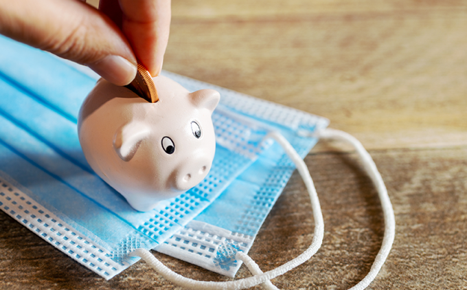 Hand puts coin in small piggy bank; rests on single-use masks on top of wood table