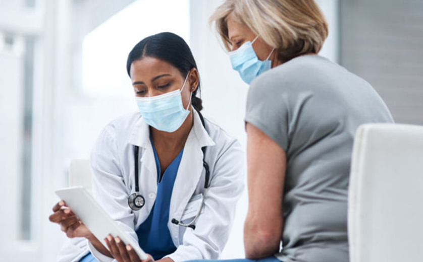 Doctor shows patient clipboard while they discuss diagnosis in hallway; seated, both masked.